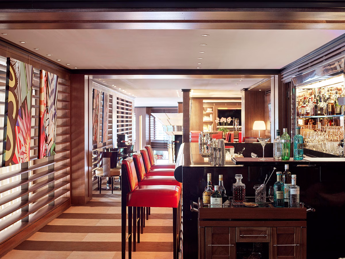 Die stilvolle BAR45 im Hotel 45 Park Lane mit einer dunklen Holztheke, leuchtend roten Barhockern, abstrakten modernen Kunstwerken an der Wand und einem hölzernen Servierwagen mit feinen Spirituosen im Vordergrund