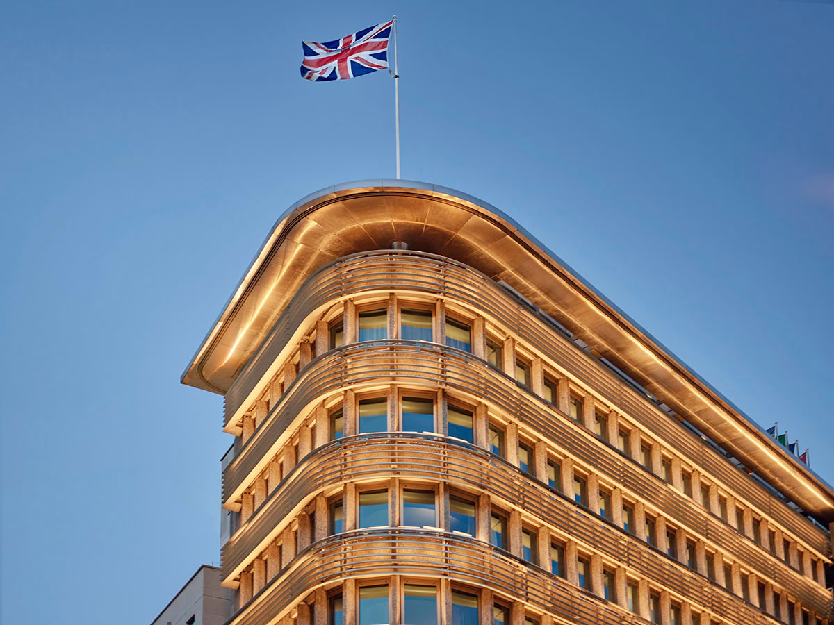 Die beleuchtete, architektonisch geschwungene Dachspitze des Hotels 45 Park Lane in der Abenddämmerung, über der stolz der britische Union Jack an einem hohen Fahnenmast vor klarem blauem Himmel weht
