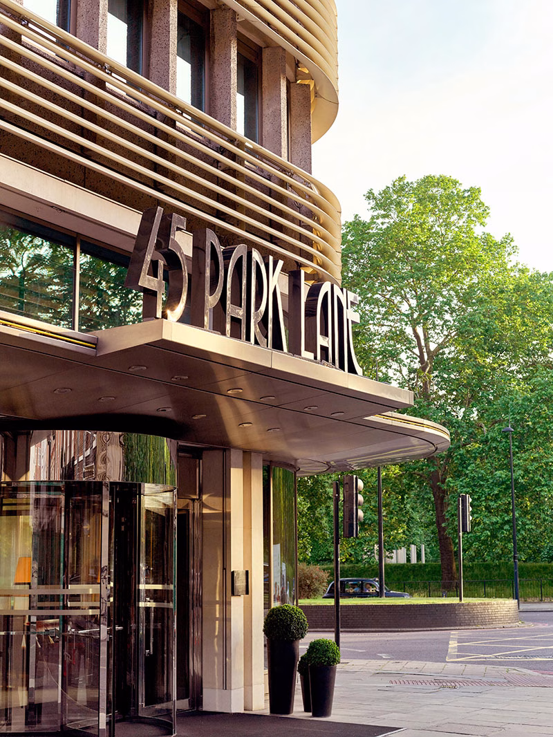Der repräsentative Eingangsbereich des Hotels 45 Park Lane mit einer gläsernen Drehtür, zwei formgeschnittenen Buchsbaumkugeln in hohen schwarzen Vasen und dem markanten, beleuchteten Schriftzug über dem Vordach