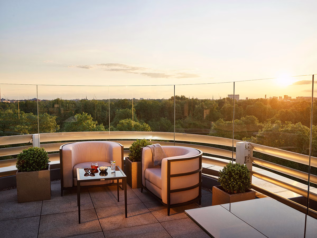 Die weitläufige Dachterrasse der Penthouse Suite im 45 Park Lane bei Sonnenuntergang mit modernen Lounge-Sesseln, einem gläsernen Couchtisch und einem Panoramablick über die Baumkronen Londons