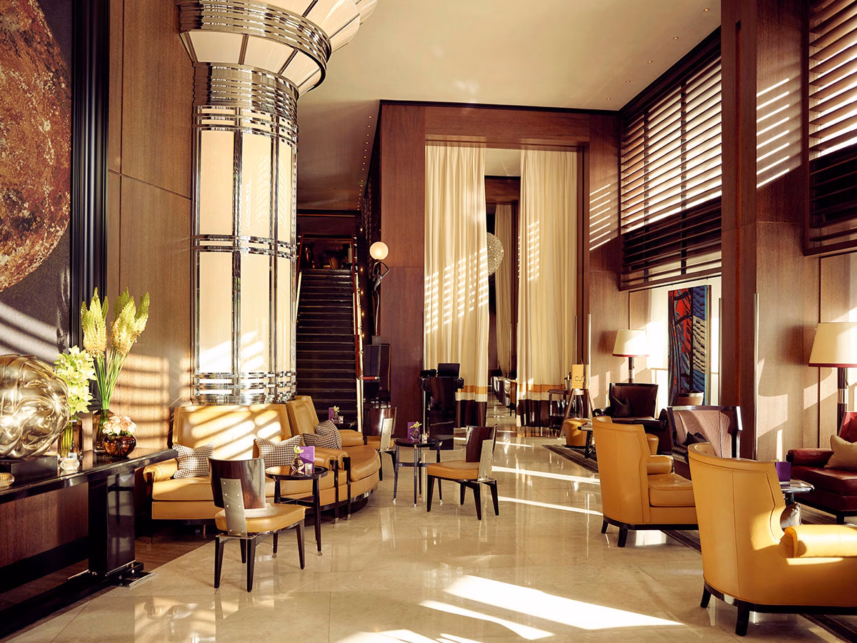 Die lichtdurchflutete Lobby des Hotels 45 Park Lane am Nachmittag mit warmen Holzwänden, eleganten gelben Ledersesseln und einer monumentalen, glänzenden Art-déco-Säule aus Chrom