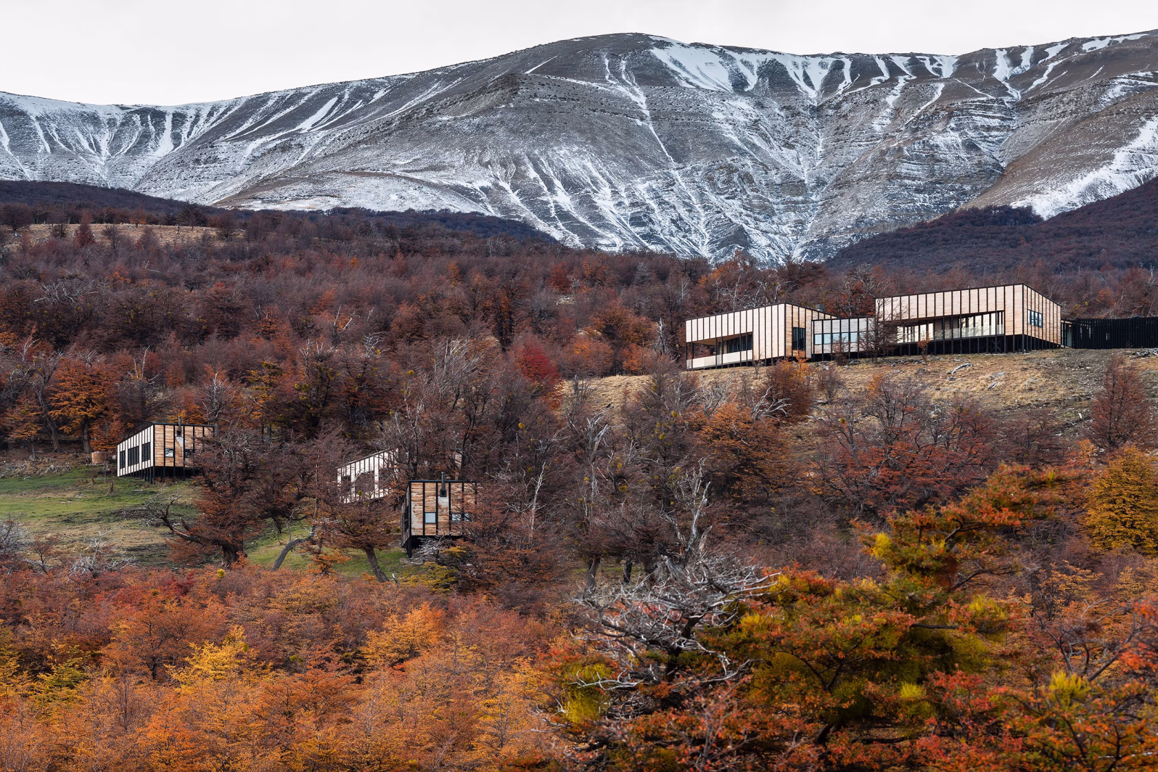 Moderne, quaderförmige Holzhütten des Awasi Patagonia auf Stelzen, eingebettet in einen herbstlich bunt gefärbten Wald vor einem schneebedeckten Bergmassiv