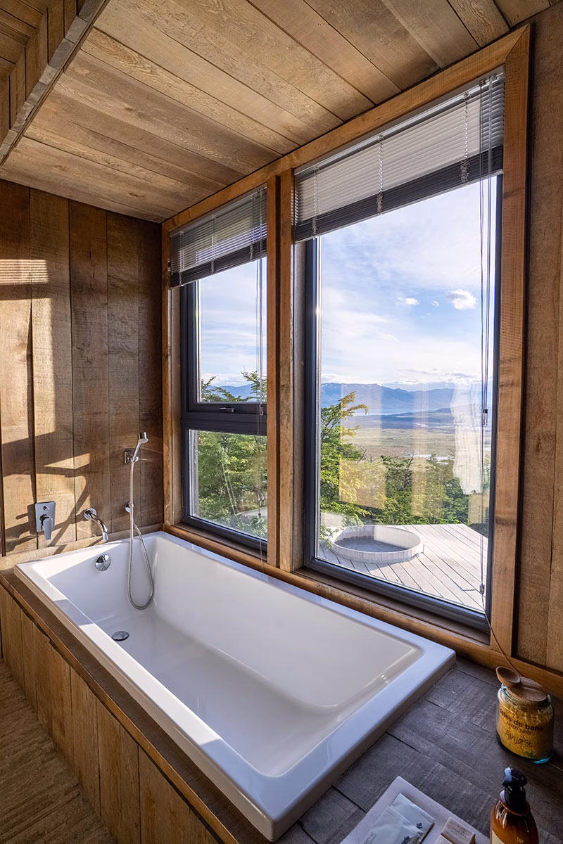 Eine in helles Holz eingelassene weiße Badewanne direkt an einem großen Eckfenster mit weitem Blick über die patagonische Landschaft und einen Whirlpool auf der Terrasse des Awasi Patagonia