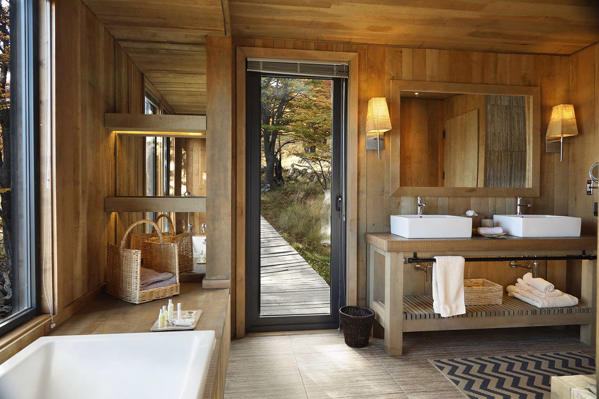 Rustikales Badezimmer im Awasi Patagonia komplett aus hellem Holz mit zwei modernen Aufsatzwaschbecken, Blick durch die geöffnete Glastür auf einen hölzernen Steg im Wald