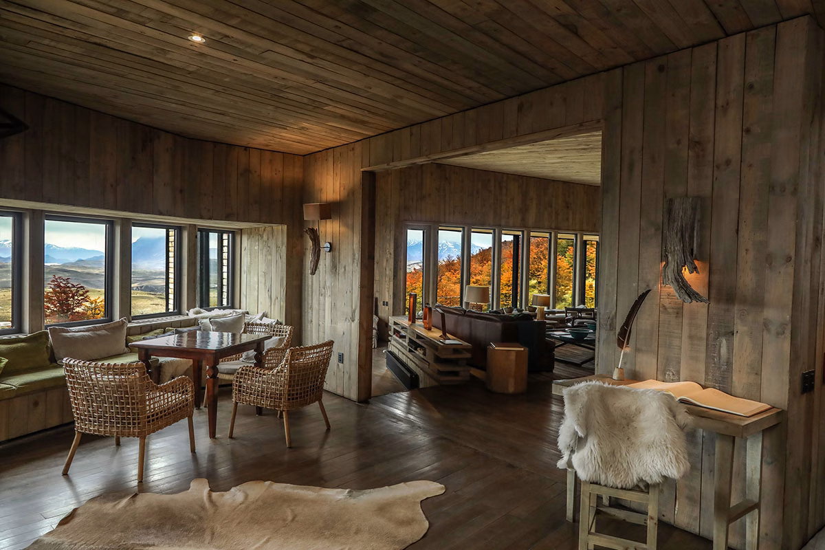 Großzügiger, komplett mit Holz verkleideter Wohnbereich im Awasi Patagonia mit Korbsesseln, einem Fellteppich und großen Panoramafenstern mit Blick auf die herbstliche Natur
