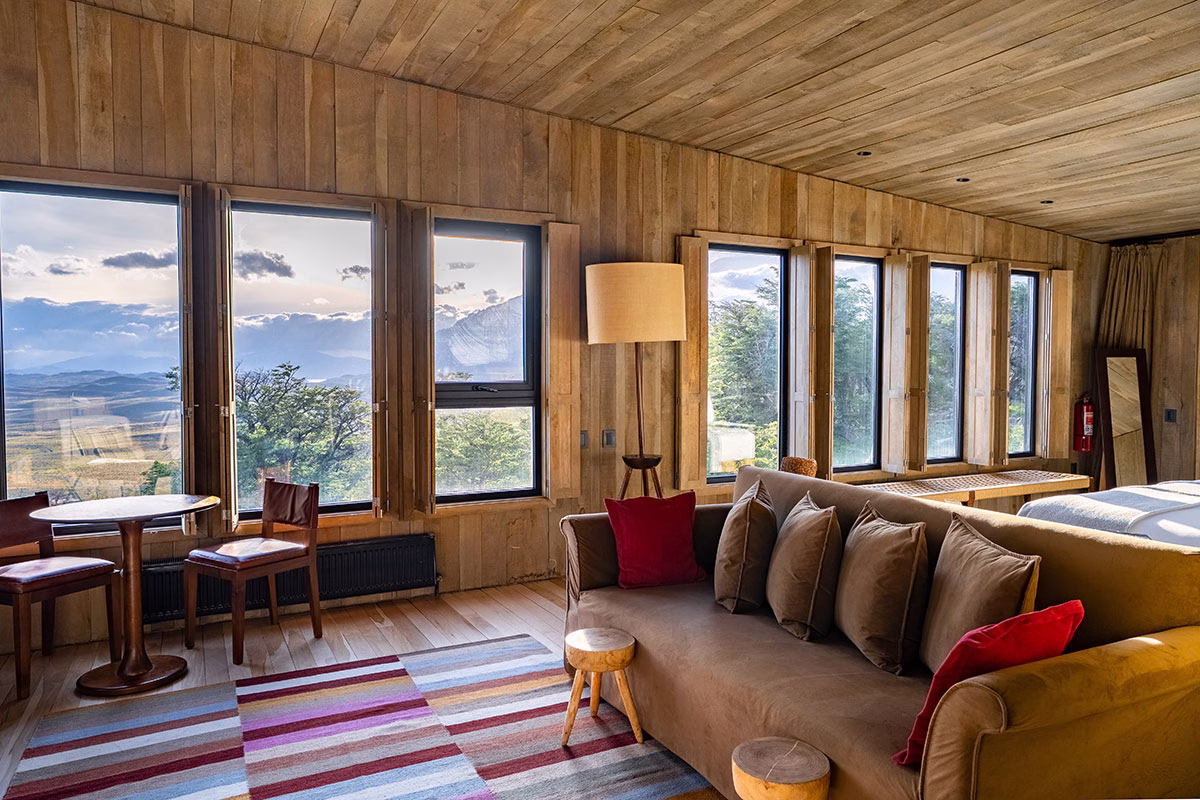Gemütlicher Wohnbereich im Awasi Patagonia mit einem braunen Sofa, bunten Kissen, einem gestreiften Teppich und einer Fensterfront, die den Blick auf die patagonische Steppe freigibt