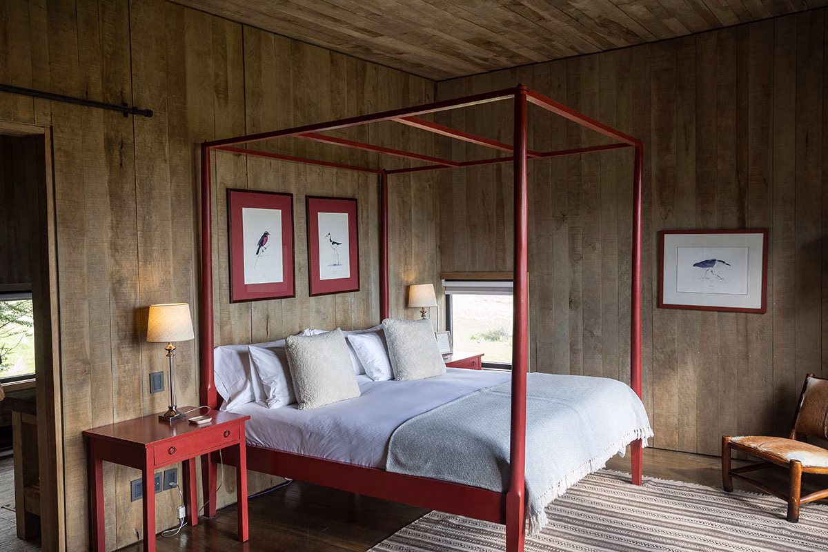 Rustikales Schlafzimmer im Awasi Patagonia mit Holzwänden, einem markanten roten Himmelbett mit weißer Bettwäsche und gerahmten Vogel-Illustrationen an der Wand