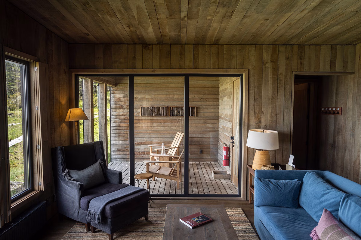 Gemütlicher Wohnbereich im Awasi Patagonia aus hellem Holz mit einem blauen Sofa und einem dunklen Sessel, Blick durch die Glasschiebetür auf eine geschützte Holzterrasse mit Liegestühlen