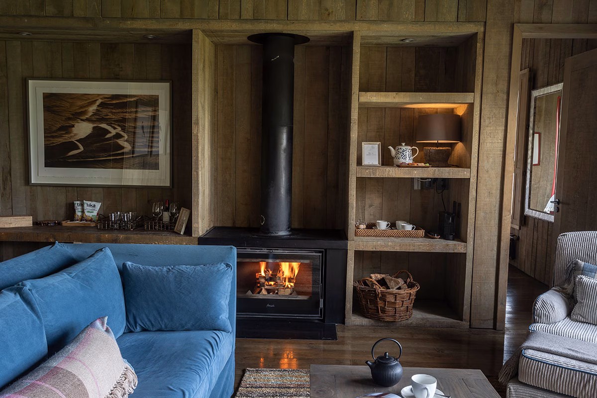 Ein brennender, moderner Kamin in einer rustikalen Holzwand im Awasi Patagonia, flankiert von einem blauen Samtsofa und Regalen mit einer Teestation