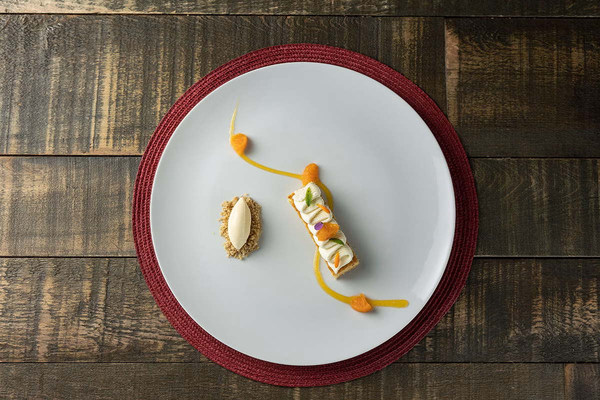Ein kunstvoll angerichtetes Dessert im Restaurant des Awasi Patagonia auf einem großen weißen Teller: Ein cremiges Törtchen mit Mandarinen-Garnitur und eine Nocke Sorbet auf Keksstreuseln, fotografiert aus der Vogelperspektive auf einem dunklen Holztisch
