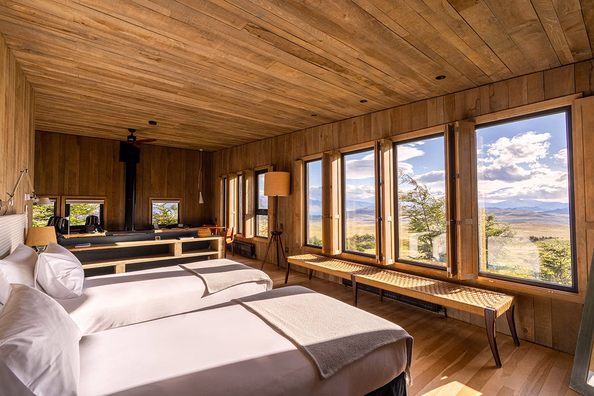 Schlafzimmer-Villa im Awasi Patagonia mit zwei nebeneinander stehenden Betten mit weißer Bettwäsche, hellen Holzwänden und einer langen Fensterfront mit weitem Blick über die Steppe unter blauem Himmel