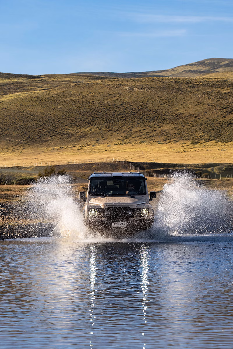 Ein beigefarbener Geländewagen des Awasi Patagonia fährt mit eingeschalteten Scheinwerfern dynamisch durch eine Flussfurt, wobei rechts und links große Wasserfontänen aufspritzen, vor einer hügeligen Landschaft