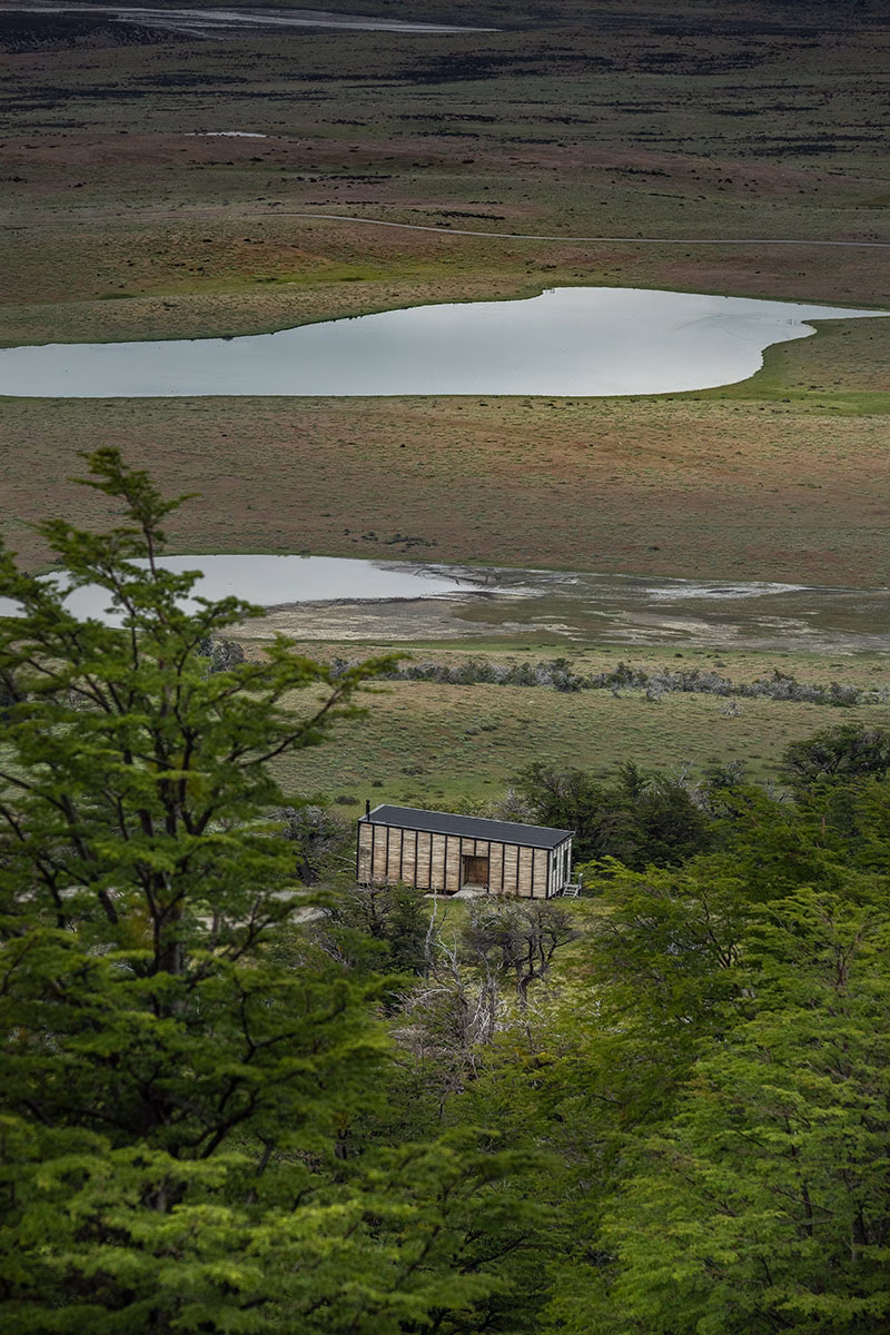 Erhöhte Ansicht einer einzelnen, rechteckigen Holzvilla des Awasi Patagonia, eingebettet in grüne Bäume und die patagonische Steppenlandschaft mit zwei flachen Seen im Hintergrund