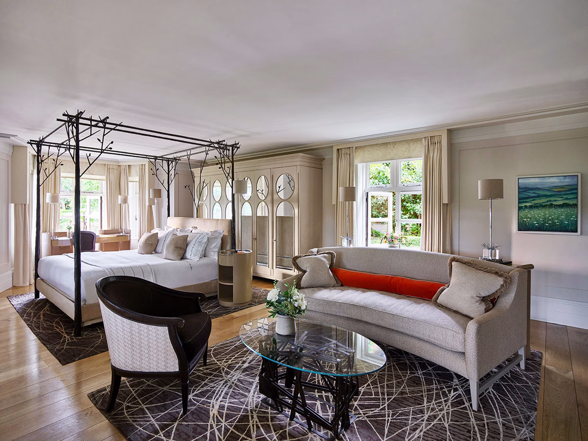 Die luxuriöse Gardener's East Cottage Suite im Coworth Park mit einem extravaganten Himmelbett aus dunklen, astähnlichen Metallstreben, einem beigen Sofa und einem gemusterten Teppich auf hellem Parkett