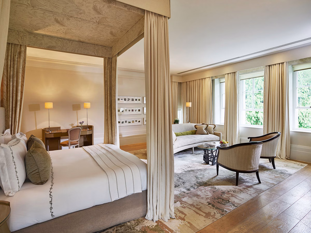 Das Schlafzimmer der Executive Weston Suite im Mansion House des Coworth Park mit einem klassischen, stoffbespannten Himmelbett, hellen Vorhängen vor großen Fenstern und einer gemütlichen Sitzecke
