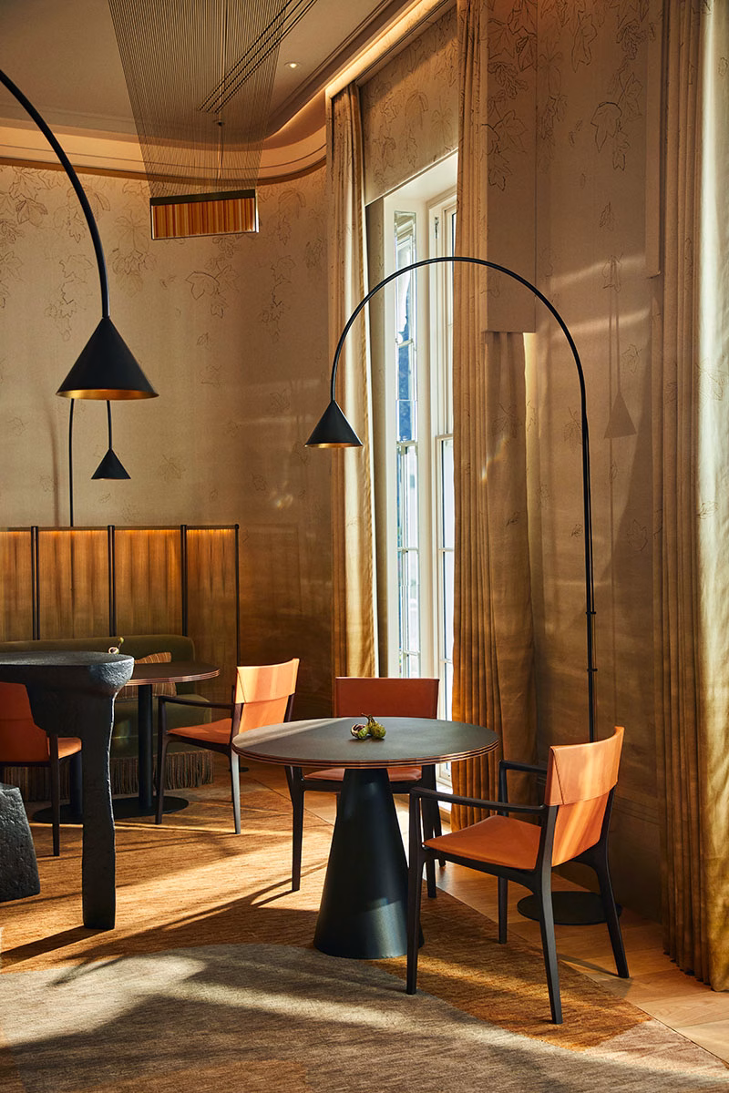 Stilvoller Essbereich im Restaurant Woven im Coworth Park mit modernen, orangefarbenen Lederstühlen, dunklen Holztischen und geschwungenen, schwarzen Stehlampen vor blattgemusterten Wänden