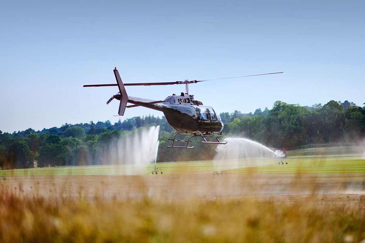 Ein eleganter schwarzer Helikopter im Tiefflug kurz vor der Landung auf dem weitläufigen Rasen des Coworth Park, im Hintergrund sind aktive Rasensprenger vor einer bewaldeten Hügelkette zu sehen