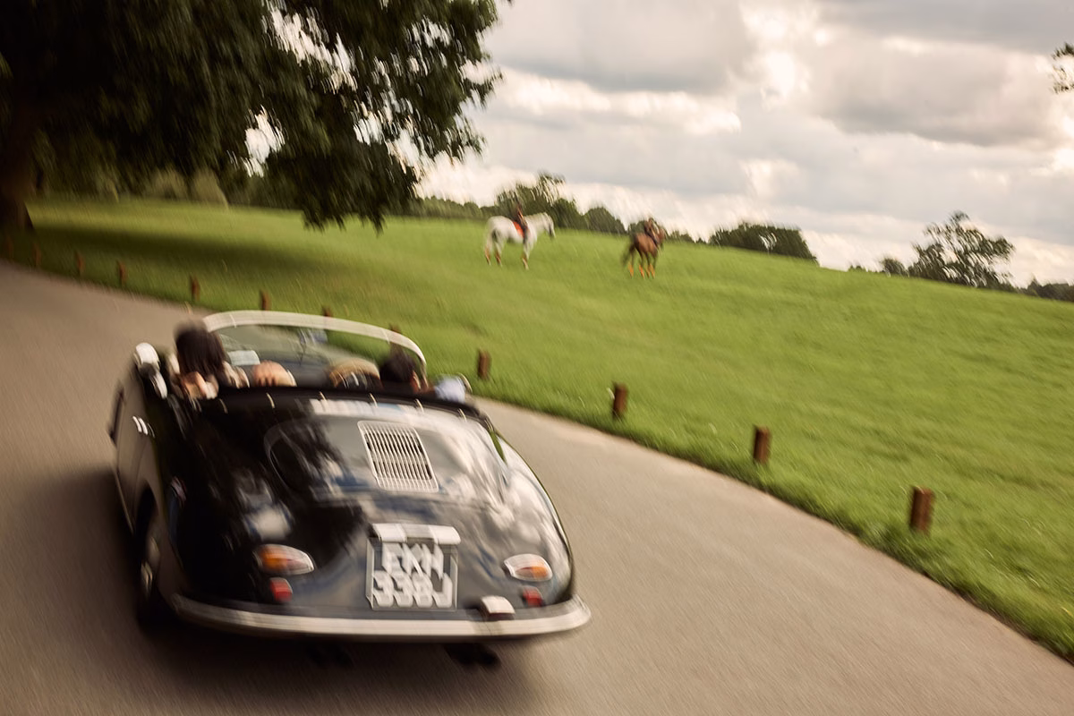 Dynamische Mitzieh-Aufnahme eines schwarzen Oldtimer-Cabrios, das mit geöffnetem Verdeck eine asphaltierte Straße entlangfährt, vorbei an einer grünen Koppel mit Pferden im Hintergrund