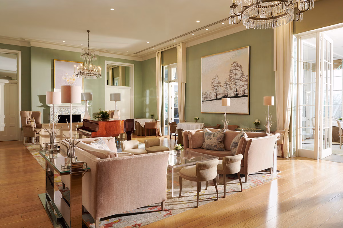 Der lichtdurchflutete, elegante Drawing Room im Coworth Park mit salbeigrünen Wänden, klassischen Kristallkronleuchtern, gemütlichen beigen Sofas und einem glänzenden Holzflügel