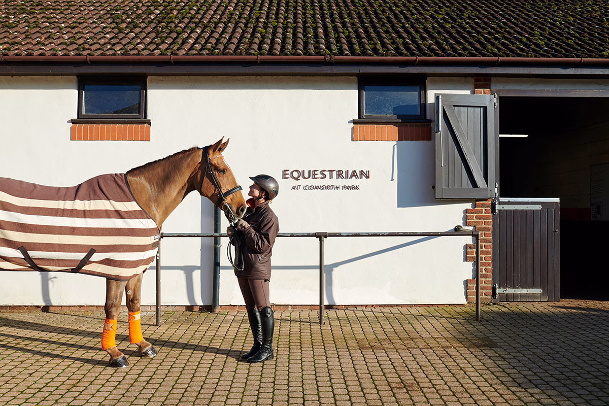 Eine Reiterin in Reitkleidung steht liebevoll blickend neben einem braunen Pferd mit gestreifter Decke vor den weißen Stallungen des „Equestrian at Coworth Park“-Zentrums