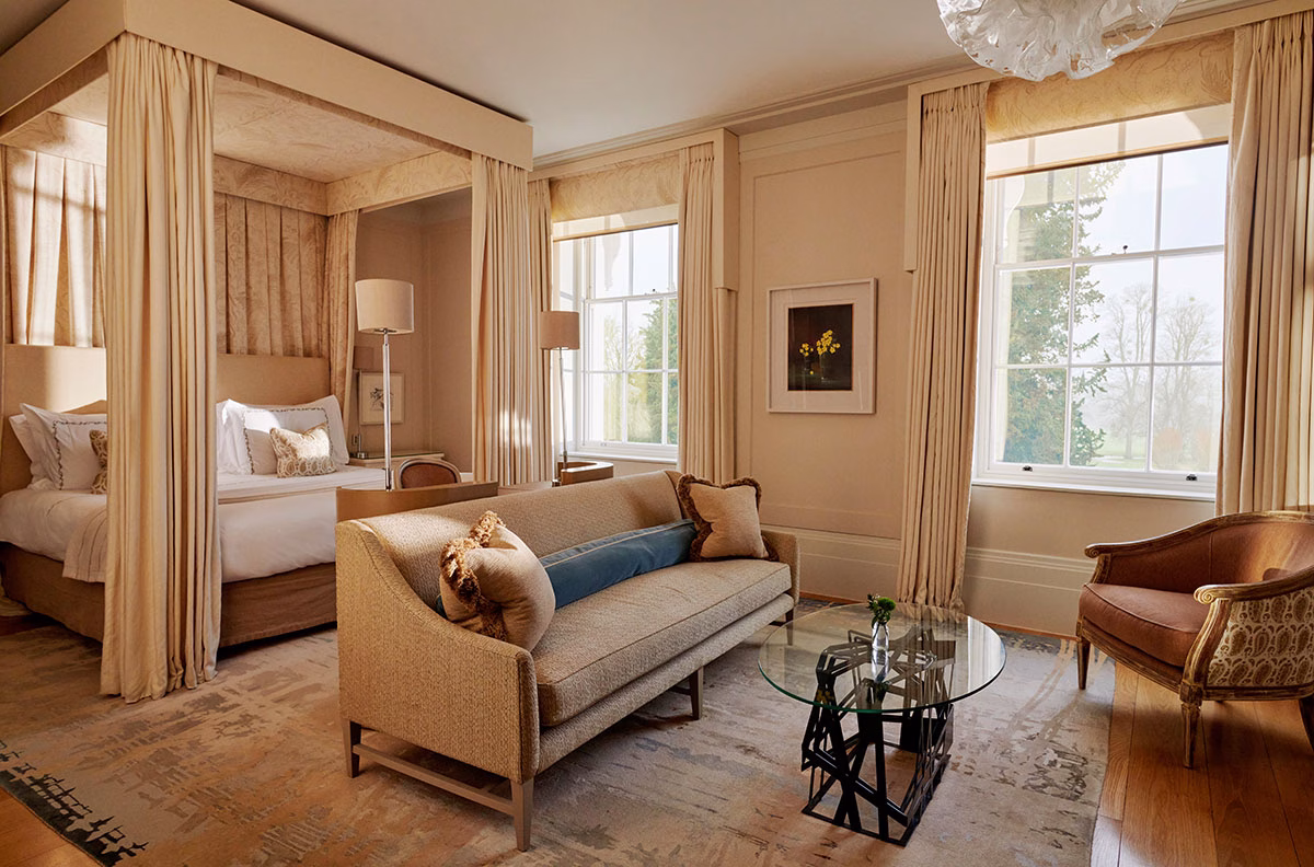 Eine helle Junior Suite im Mansion House des Coworth Park mit einem edlen Himmelbett, einem eleganten beigen Sofa, warmen Holztönen und großen Fenstern mit Blick in den grünen Hotelgarten