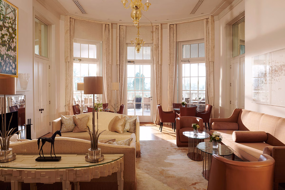 Der lichtdurchflutete, halbkreisförmige Summer Room im Coworth Park mit bodentiefen Fenstertüren zur Terrasse, eleganten cremefarbenen Sofas und edlen Kronleuchtern