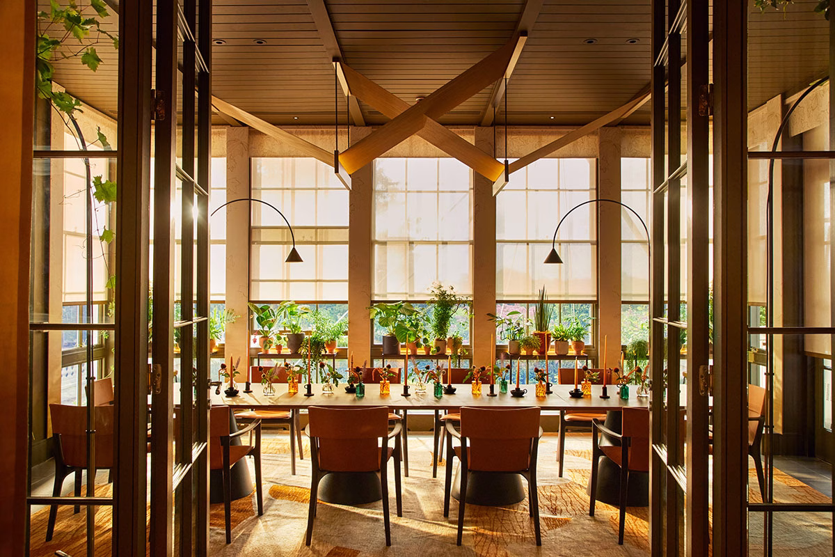 Blick durch eine geöffnete Glastür in den Wintergarten des Restaurants Woven by Adam Smith, mit einer langen, herbstlich dekorierten Kaffeetafel und einer kunstvollen Holzskulptur an der Decke