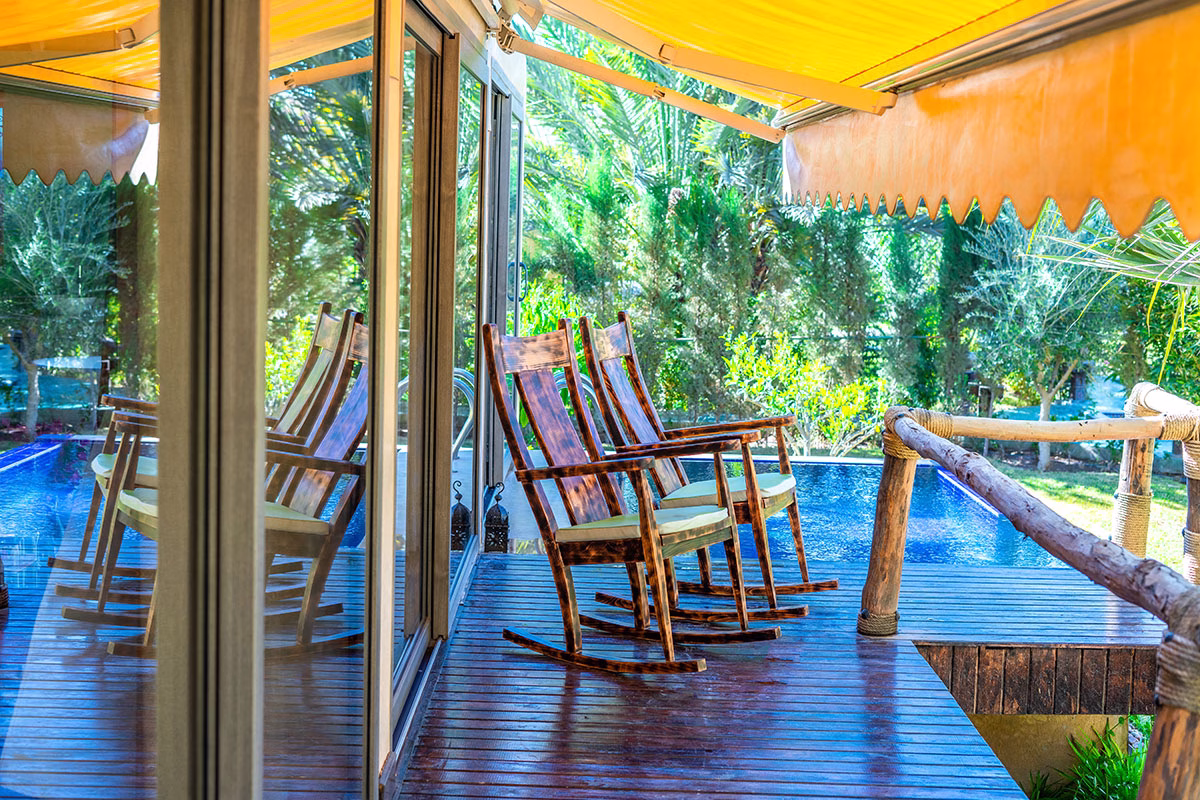 Holzterrasse eines Bungalows im Dar Hnini mit zwei Schaukelstühlen aus dunklem Holz, einer gelben Markise und direktem Blick auf das blaue Wasser des privaten Swimmingpools
