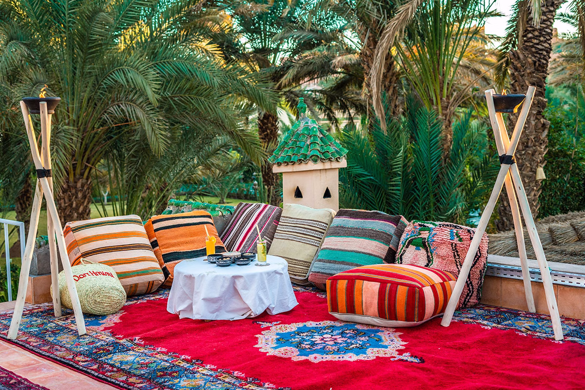 Traditionell marokkanischer Loungebereich auf einer Dachterrasse des Dar Hnini mit bunten orientalischen Sitzkissen und Poufs auf einem roten Teppich, flankiert von Fackeln vor einer Palmenkulisse