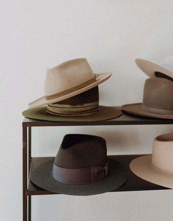 Hochwertige, stilvolle Filzhüte in verschiedenen Natur- und Erdtönen wie Beige, Olivgrün und Braun, ordentlich aufgereiht auf den dunklen Metallböden eines minimalistischen Regals vor einer hellen Wand