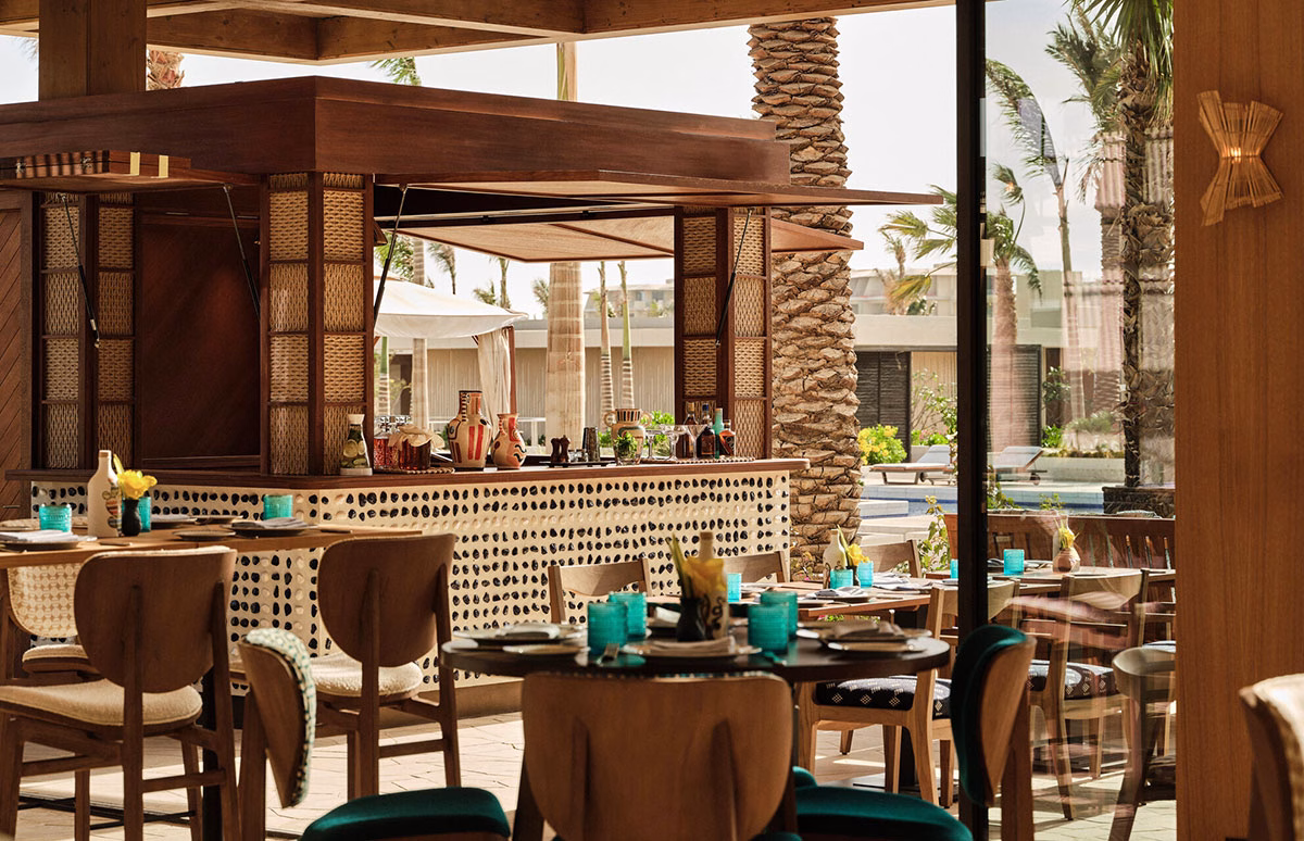Eine einladende, mit Mosaiksteinen verzierte Open-Air-Bar im Four Seasons AMAALA mit Blick auf Restauranttische, Holzstühle und den angrenzenden Außenpool