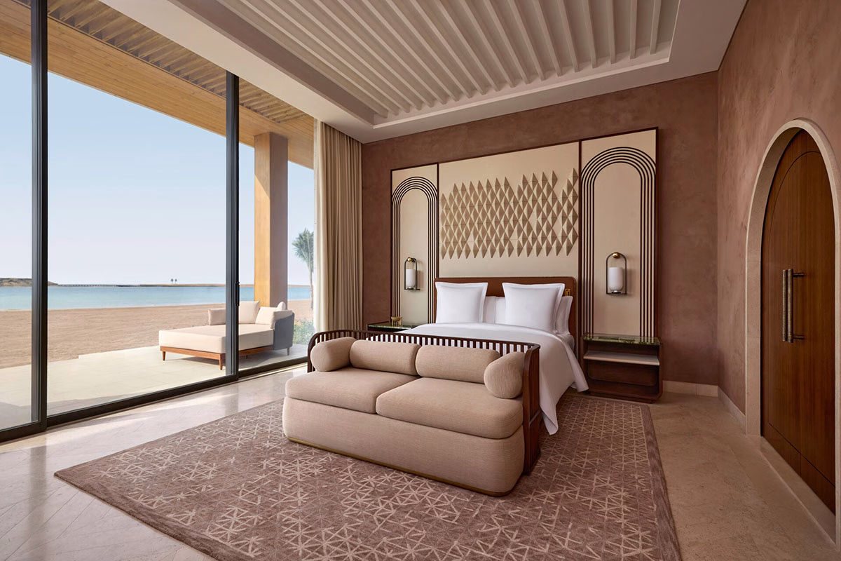 Elegantes Hotelzimmer im Four Seasons AMAALA mit einem Doppelbett, einem davorstehenden Sofa und einer raumhohen Glasschiebetür, die direkt auf die Terrasse am Sandstrand führt
