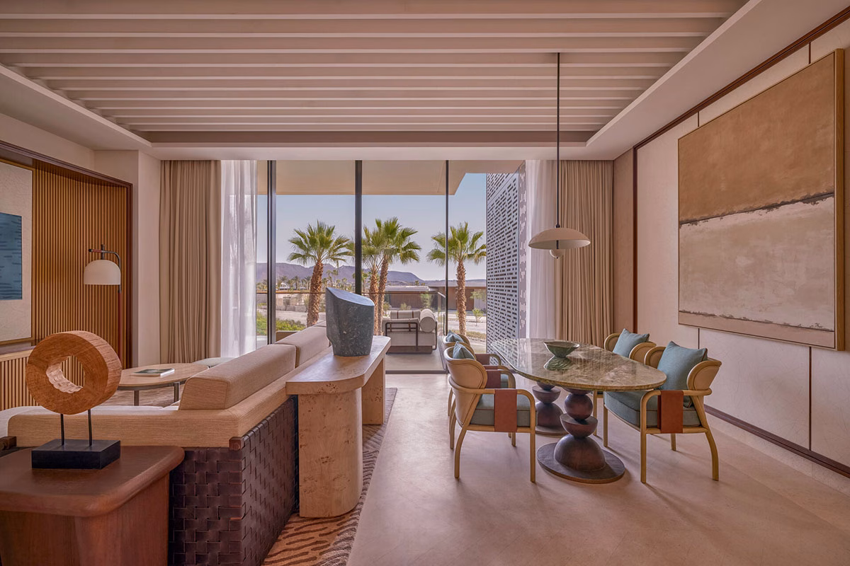 Lichtdurchfluteter Wohn- und Essbereich einer Suite im Four Seasons AMAALA mit einem eleganten Steintisch, modernen Designermöbeln und Kunstwerken sowie einer Fensterfront mit Blick auf Palmen