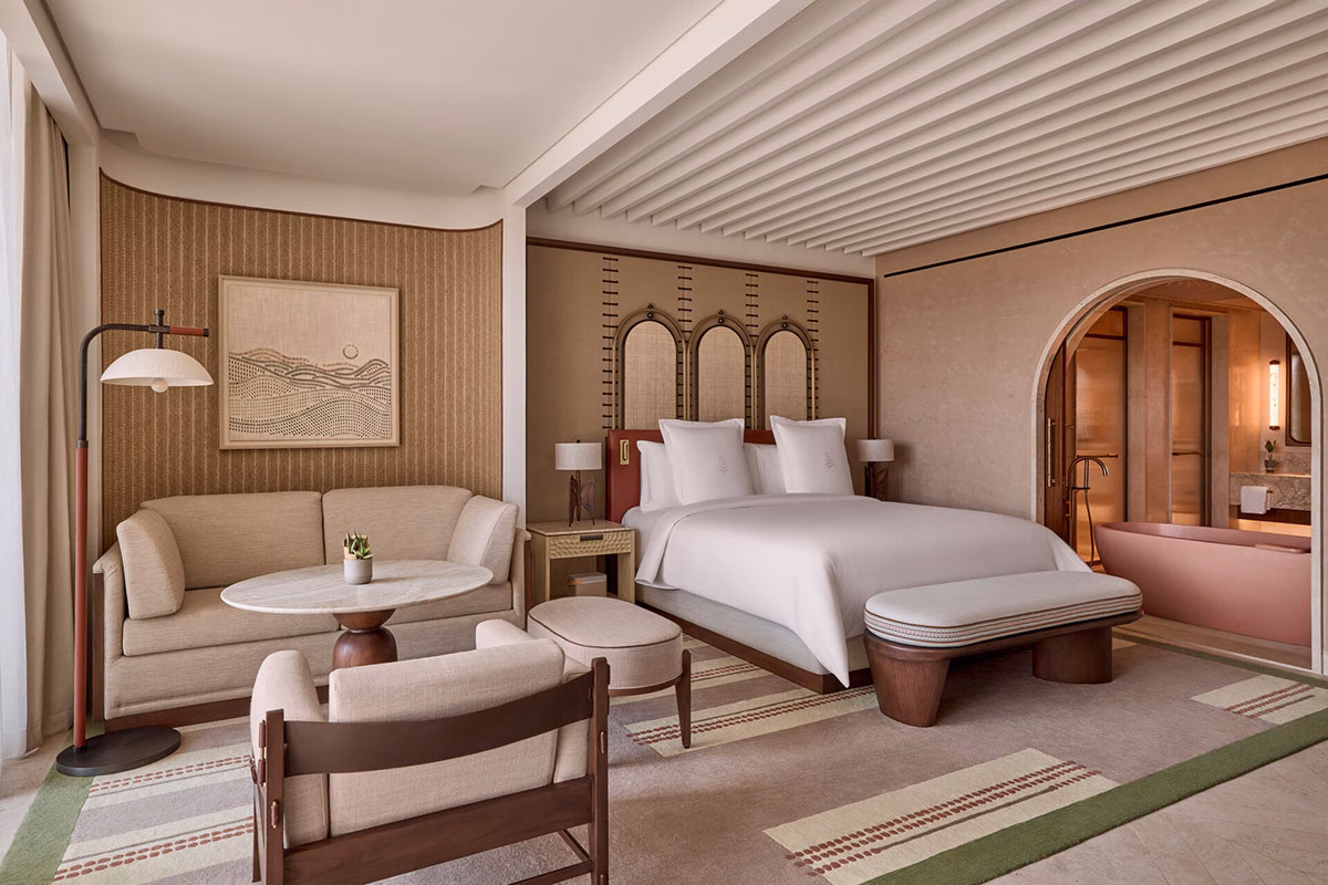 Modernes Schlafzimmer im Four Seasons AMAALA in warmen Erdtönen mit integriertem Wohnbereich, Sofa, Sessel und Blick auf den angrenzenden Badezimmerbereich mit rötlicher Badewanne