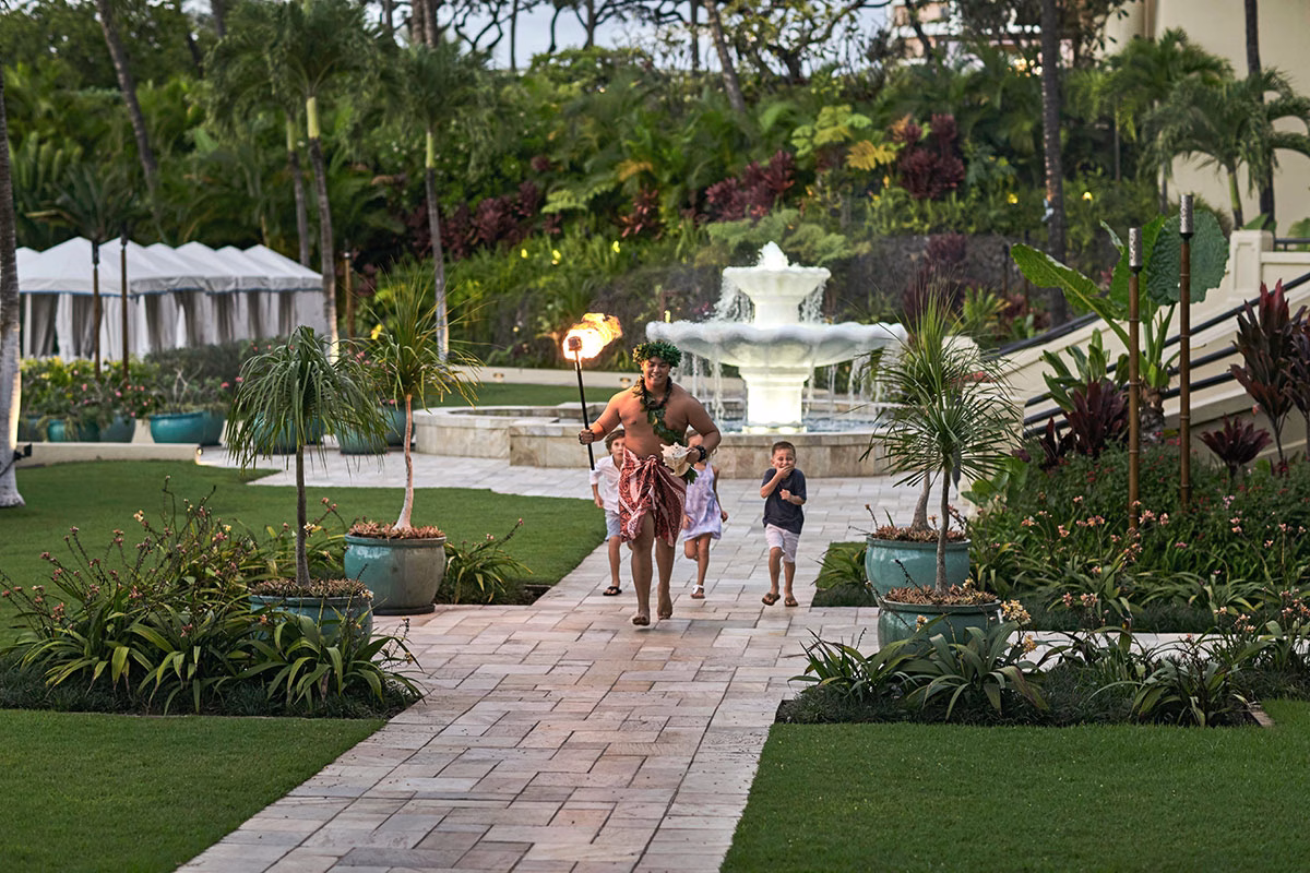 Ein hawaiianischer Fackelträger läuft im Garten des Four Seasons Maui at Wailea an einem beleuchteten Springbrunnen vorbei, gefolgt von fröhlichen Kindern