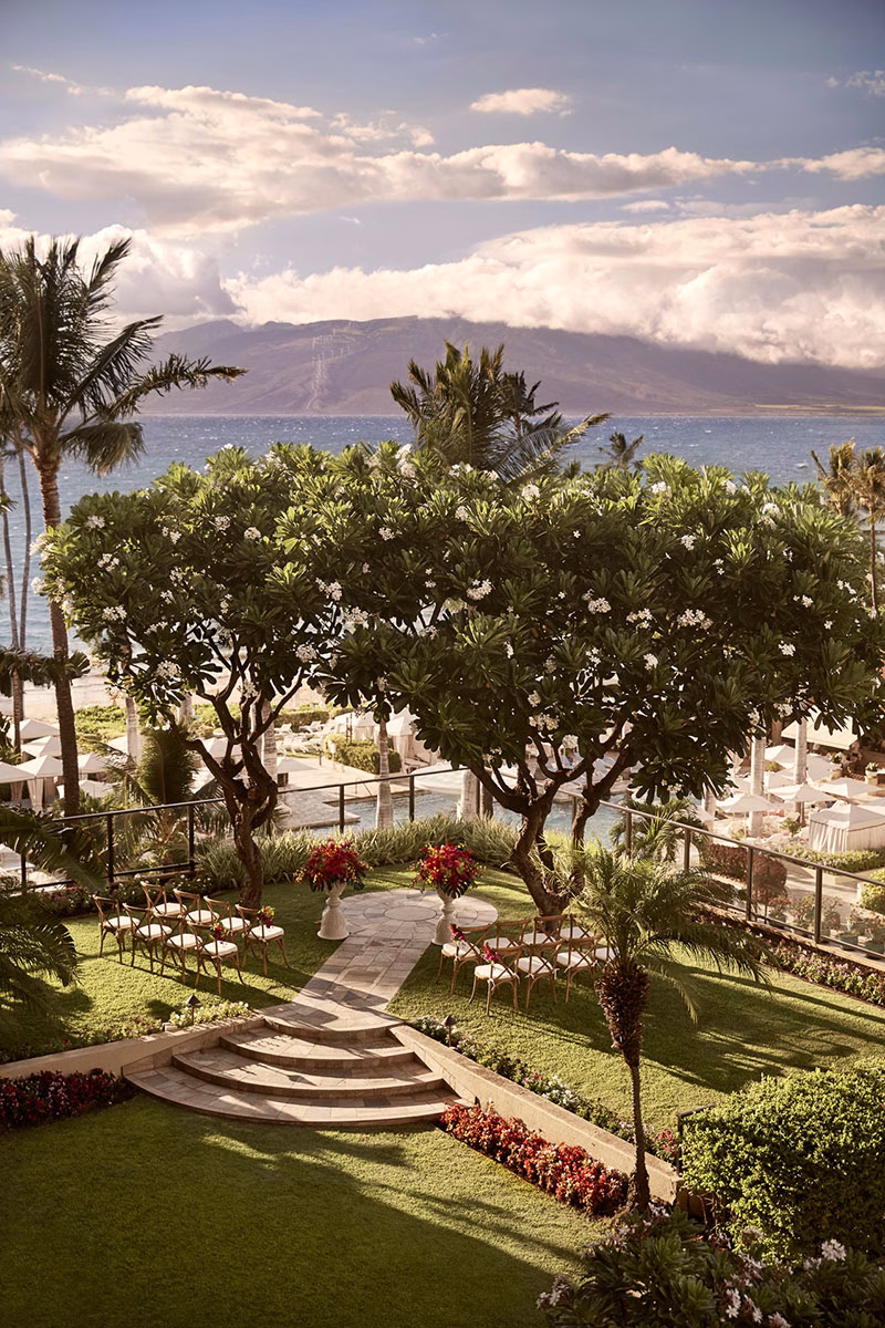 Ein idyllischer Hochzeits- oder Eventplatz im Garten des Four Seasons Maui at Wailea, eingerahmt von blühenden Bäumen und weißen Stühlen mit Blick auf das Meer