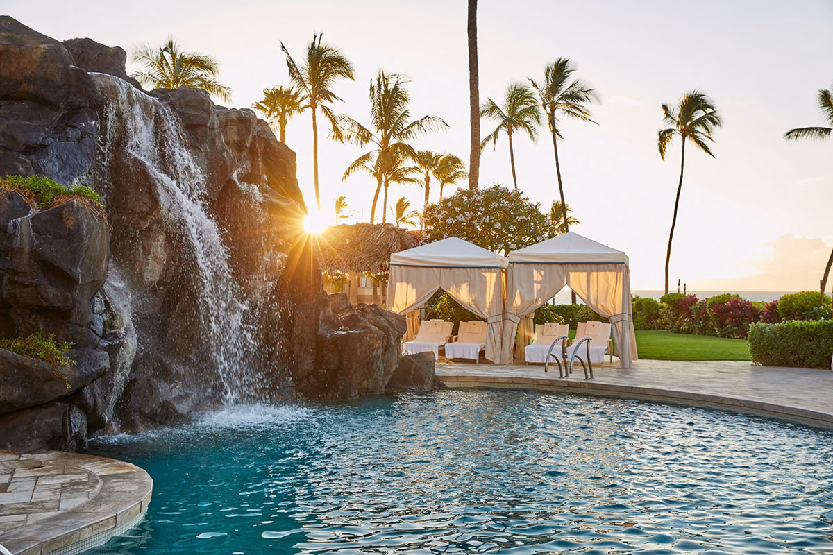 Ein künstlicher Wasserfall stürzt über dunkle Felsen in den Pool des Four Seasons Maui at Wailea, im Hintergrund glänzen weiße Cabanas in der warmen Abendsonne