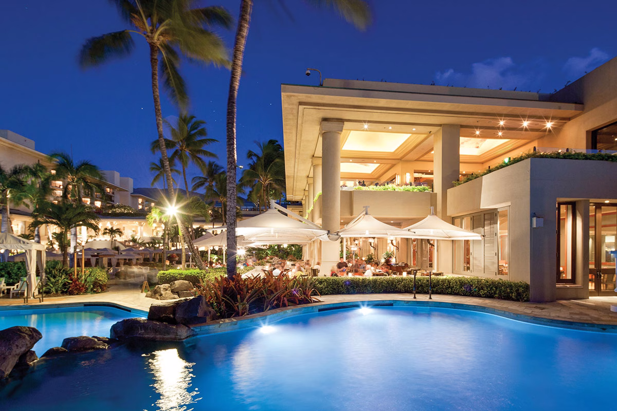 Der stimmungsvoll beleuchtete Hauptpool des Four Seasons Maui at Wailea bei Nacht, gesäumt von Palmen und der hell erleuchteten Hotelarchitektur im Hintergrund