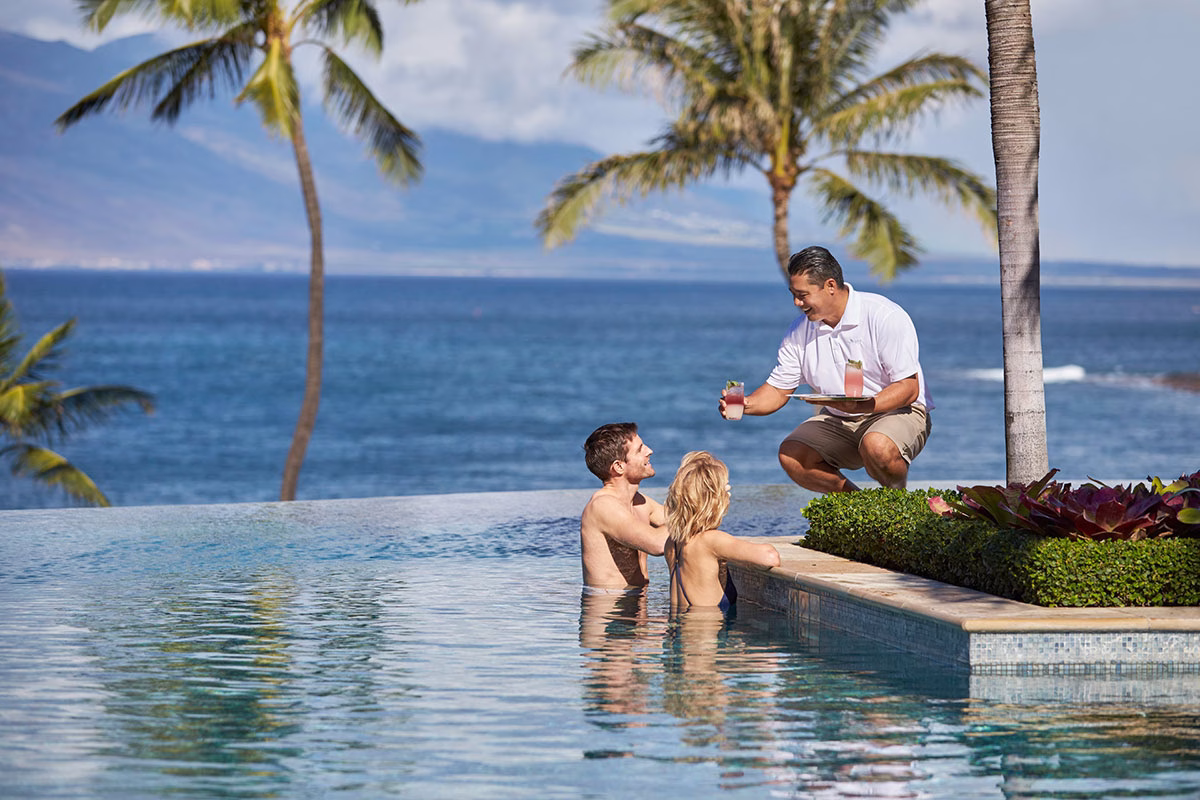 Ein Paar entspannt im Infinity-Pool des Four Seasons Maui at Wailea, während ein lächelnder Mitarbeiter erfrischende Cocktails auf einem Tablett serviert, mit dem Pazifik im Hintergrund