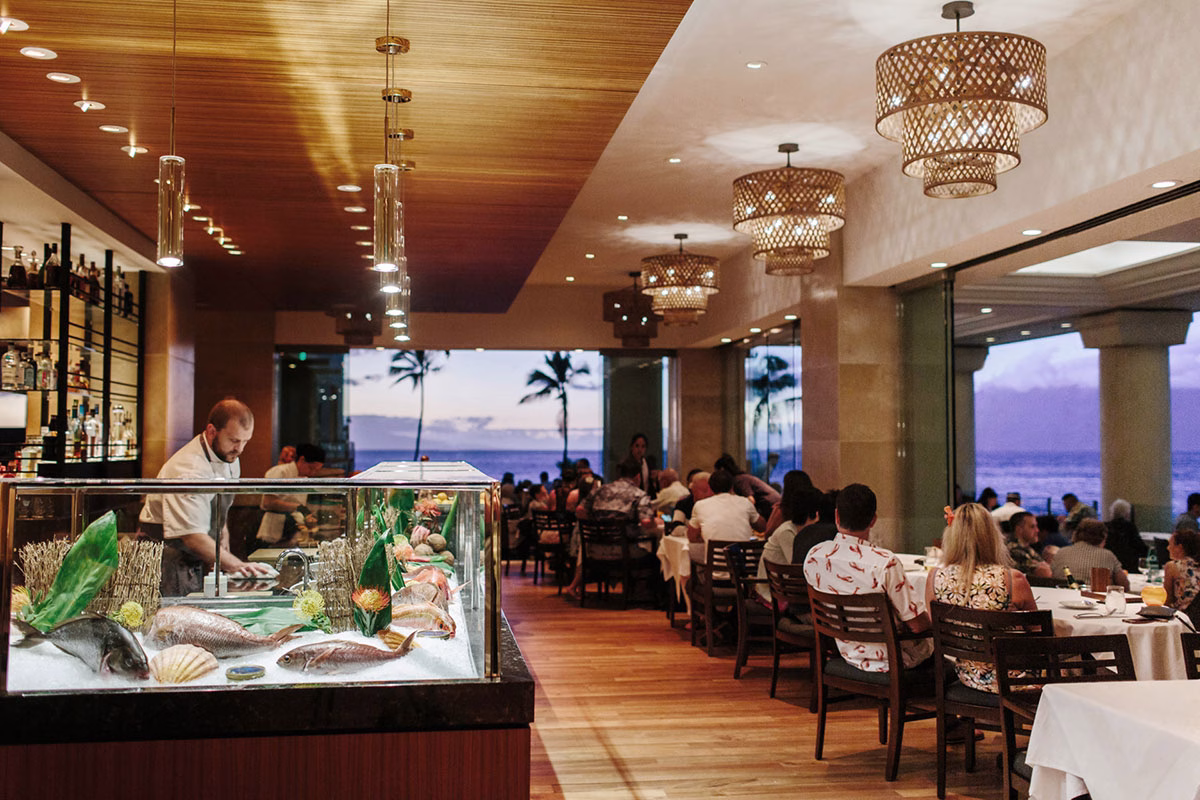 Das stilvolle Innenrestaurant im Four Seasons Maui at Wailea mit einer Auslage für frischen Fisch, gemütlichen Tischen und einer offenen Front mit Blick auf das abendliche Meer
