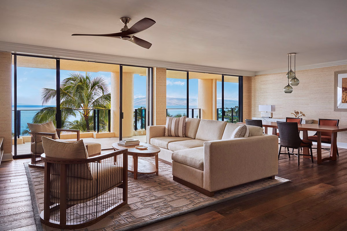 Das luxuriöse Wohnzimmer einer weitläufigen Suite im Four Seasons Maui at Wailea mit einer beigen Coucheincke, edlem Holzfußboden und einer Fensterwand, die sich komplett zum Meer hin öffnen lässt