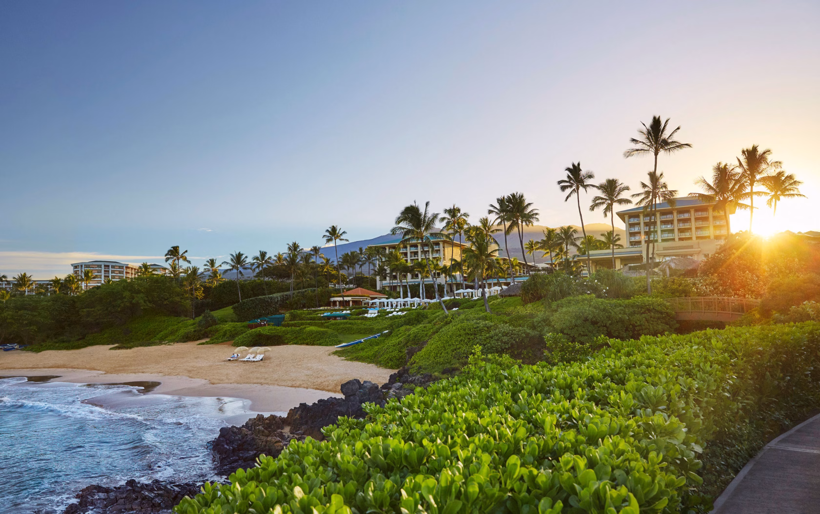 Die Silhouette des Four Seasons Maui at Wailea Resorts an einem palmengesäumten Sandstrand, während die Sonne flach über dem Horizont steht und die Küste in goldenes Licht taucht