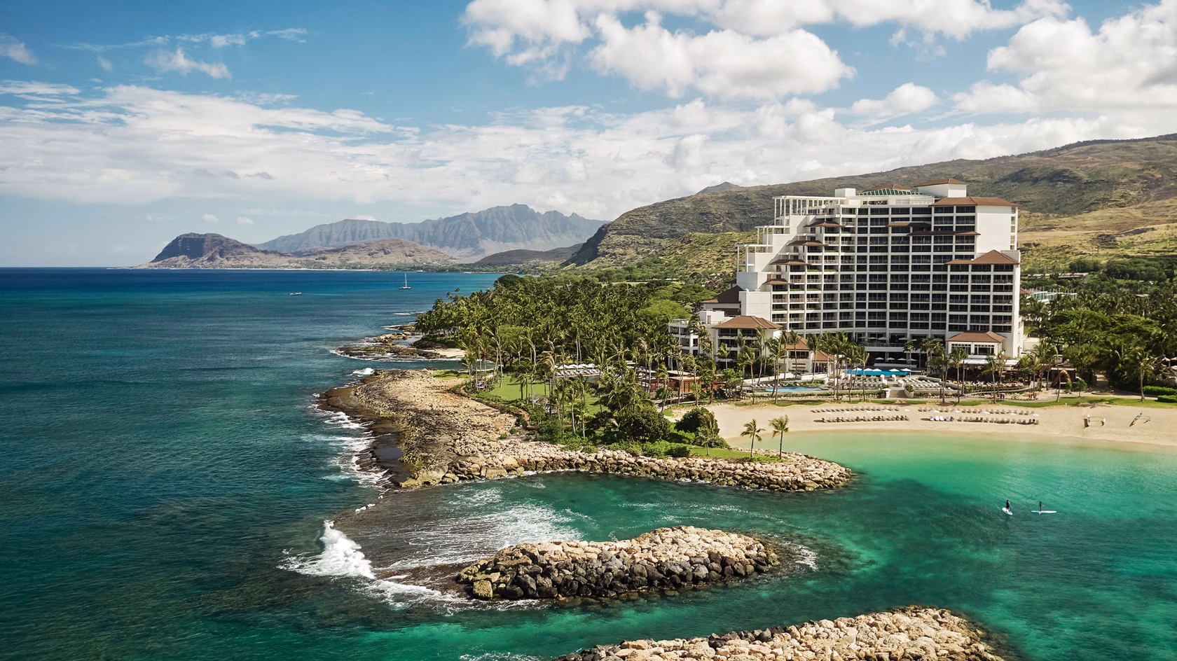 Das markante, weiße Hauptgebäude des Four Seasons Oahu an einer geschützten Badebucht; im Hintergrund erstreckt sich die weite, bergige Küstenlandschaft von Hawaii unter leicht bewölktem Himmel