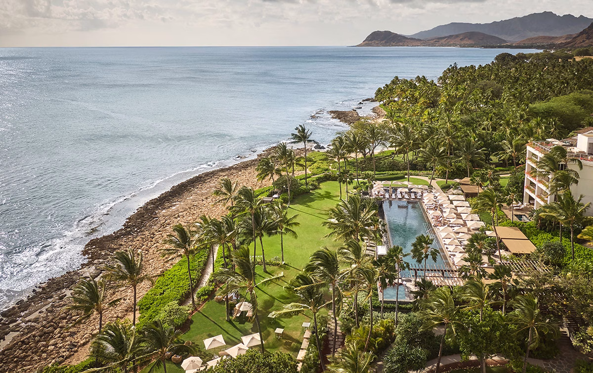 Luftaufnahme des eleganten, von Palmen gesäumten Infinity-Pools für Erwachsene im Four Seasons Oahu mit zahlreichen weißen Sonnenschirmen auf einer grünen Rasenfläche direkt an der felsigen Küste