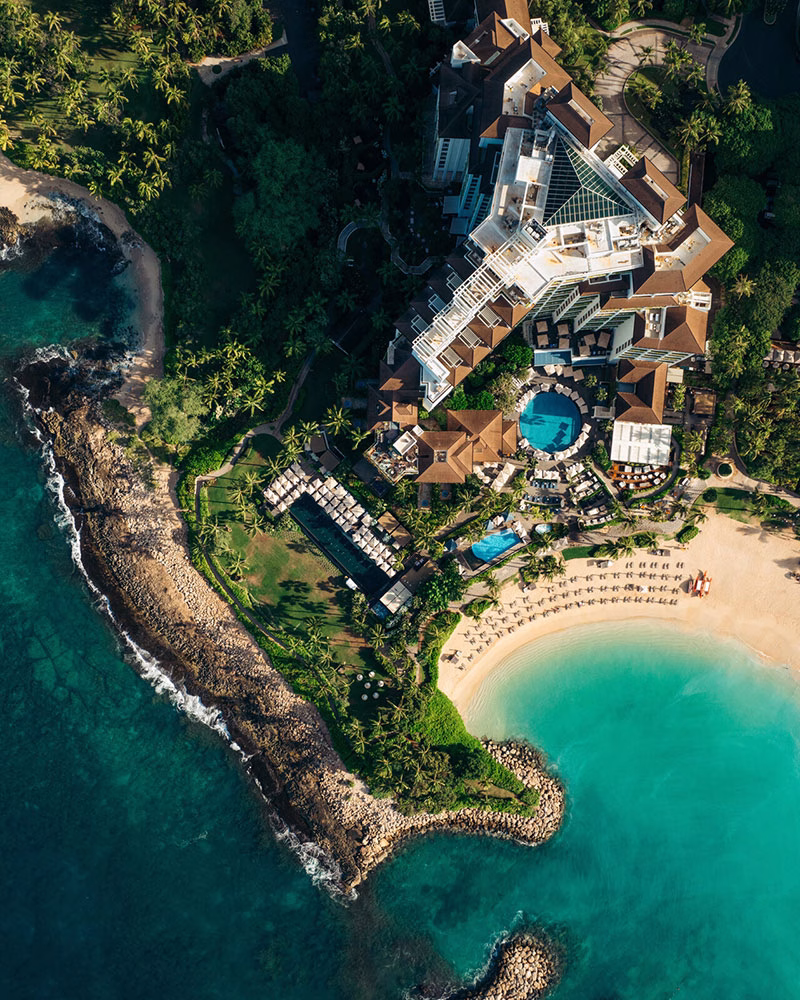 Spektakulärer Blick gerade nach unten auf die geometrische Architektur des Hotels Four Seasons Oahu, die blauen Poolbecken und den sichelförmigen Badestrand im warmen Sonnenlicht