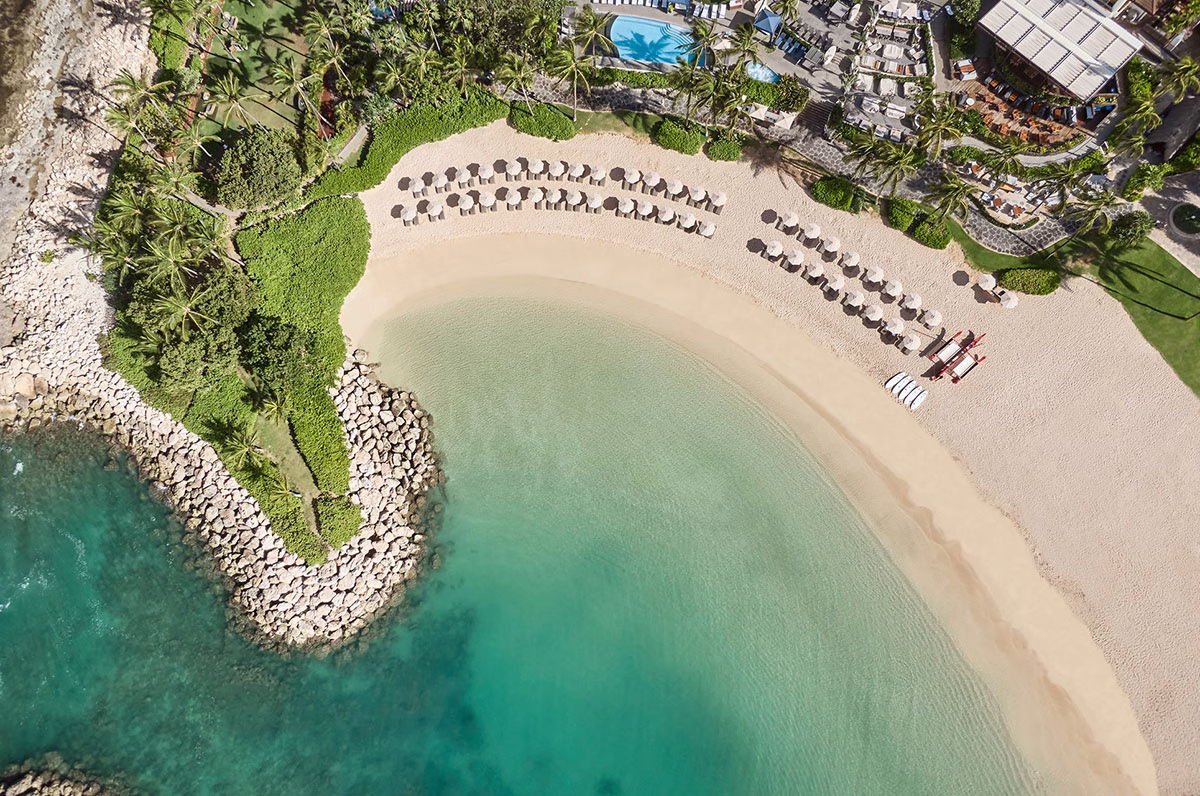 Senkrechte Vogelperspektive auf den perfekt geschwungenen, weißen Sandstrand des Four Seasons Oahu mit ordentlich aufgereihten Sonnenschirmen und kristallklarem, ruhigem Wasser an einer Steinklippe