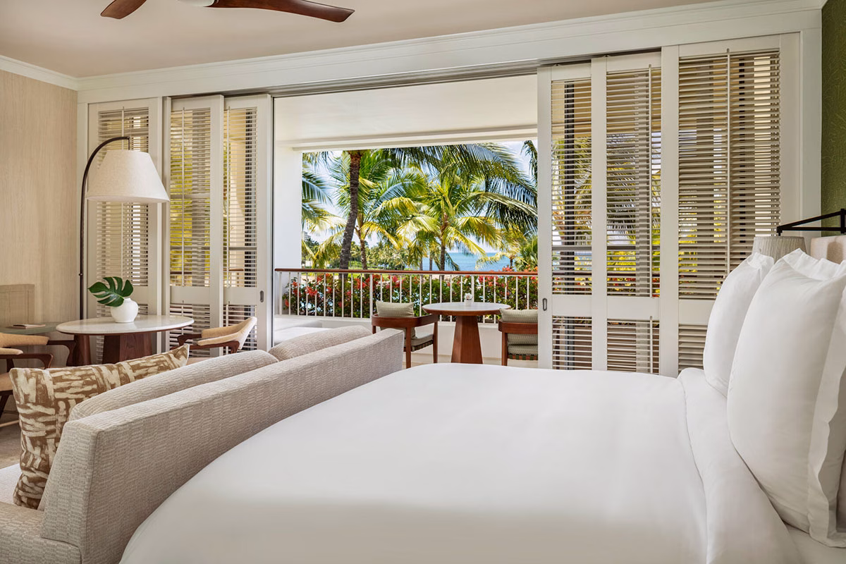 Helles, luxuriöses Hotelzimmer im Four Seasons Oahu mit einem großen weißen Bett und geöffneten Schiebetüren aus Holz-Lamellen, die den Blick auf einen Balkon mit Palmen und das Meer freigeben