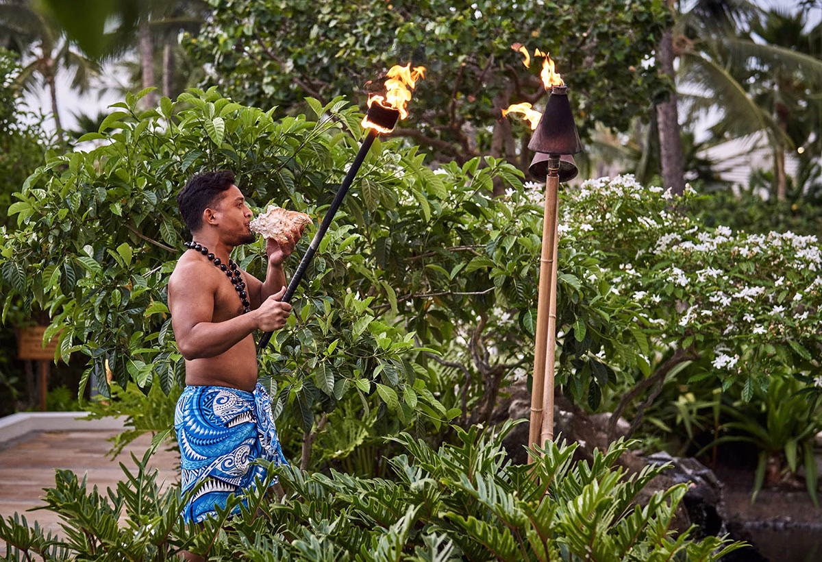 Ein hawaiianischer Mann in traditioneller, blau gemusterter Kleidung bläst bei einer Kultur-Vorführung im Four Seasons Oahu vor brennenden Tiki-Fackeln in eine große Seemuschel