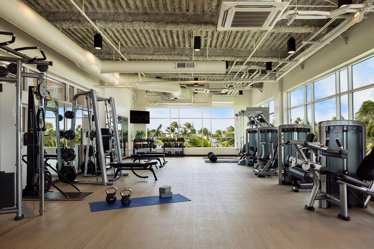 Das moderne Fitnessstudio im Four Seasons Oahu mit Kraft- und Ausdauergeräten sowie einer großen Fensterfront, die den Blick auf Palmen und das Meer freigibt
