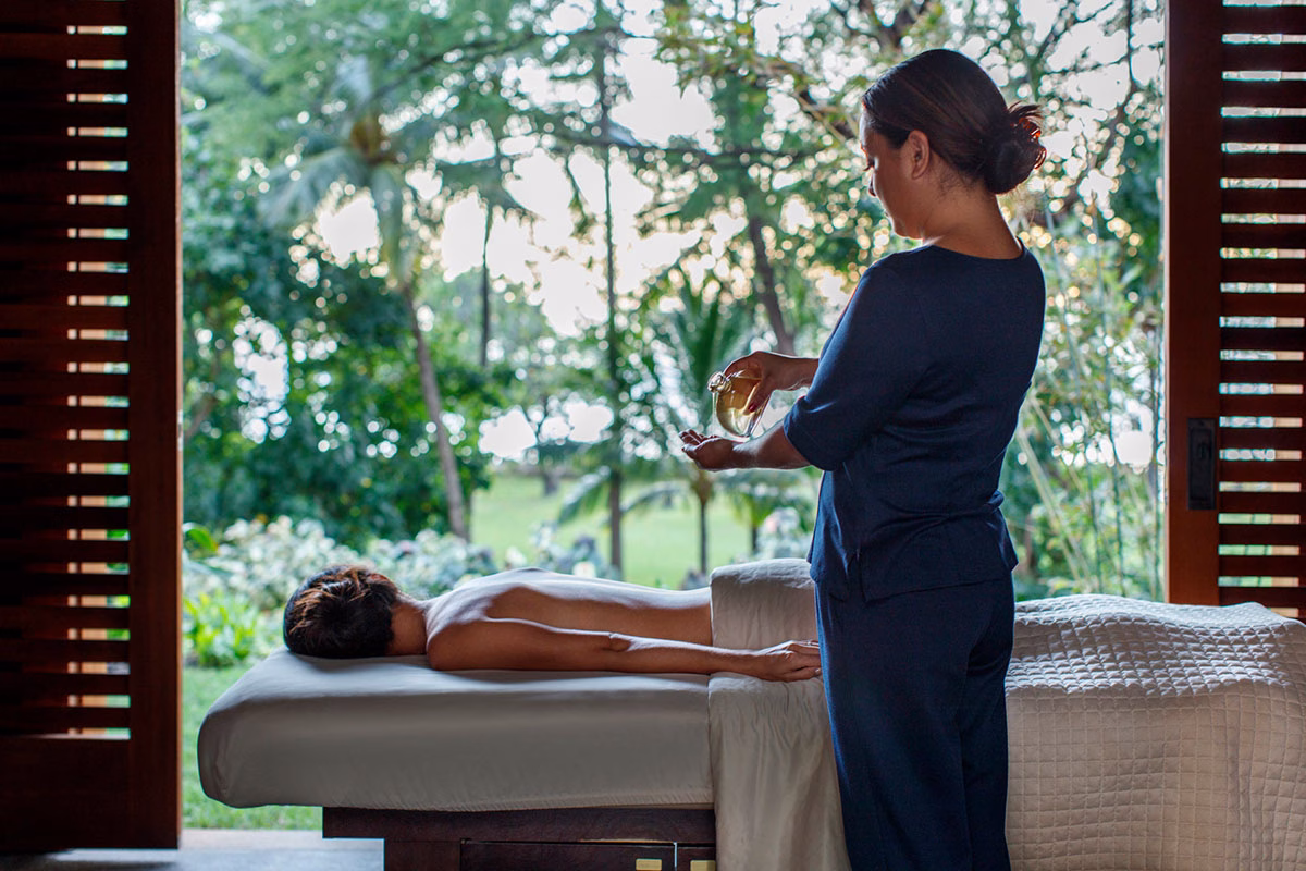 Eine Wellness-Behandlung im Spa des Four Seasons Oahu, bei der eine Therapeutin Massageöl auf den Rücken einer entspannten Frau aufträgt, mit Blick in den tropischen Garten